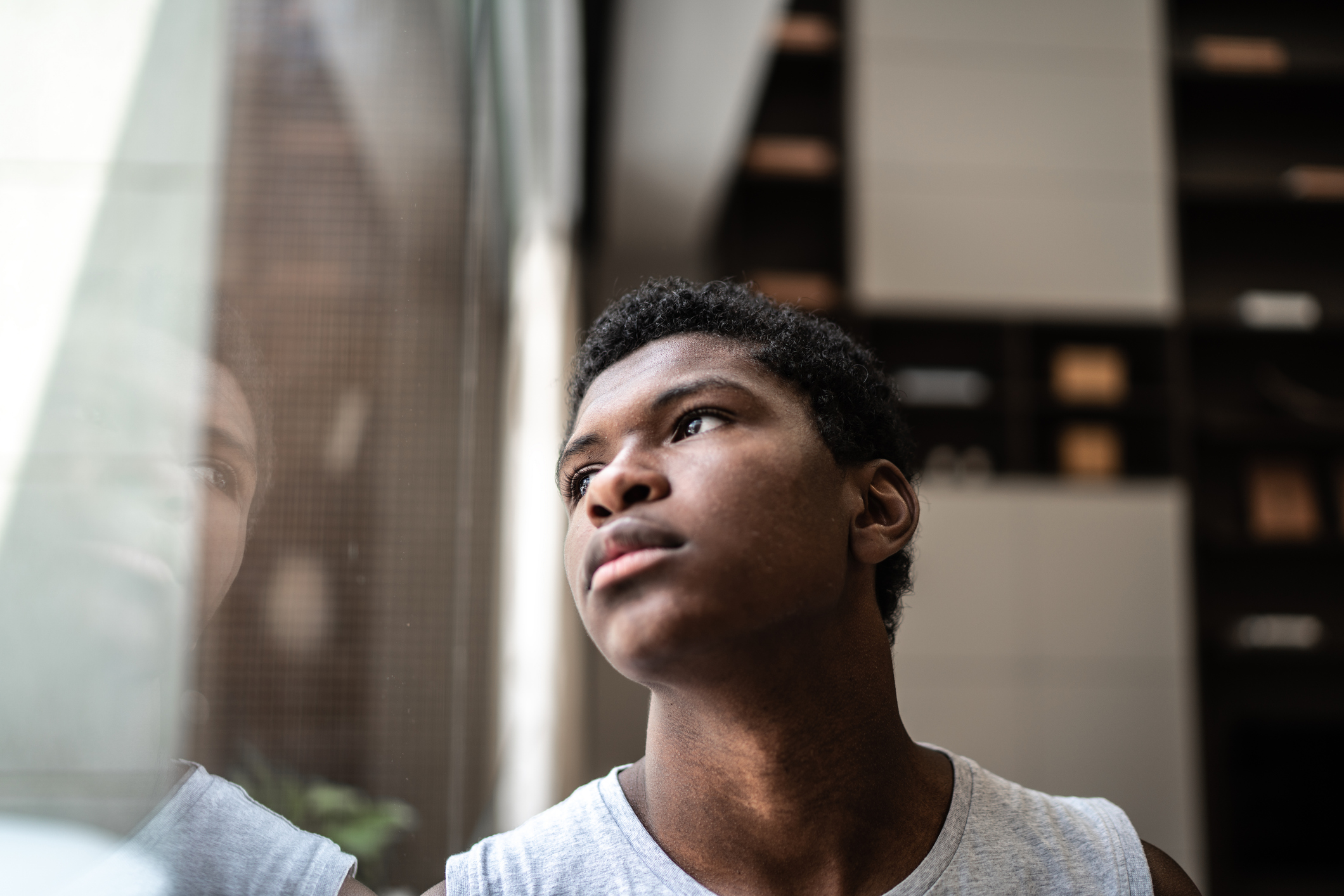 A Black teen boy gazes out a window with a quiet expression, his reflection visible beside him